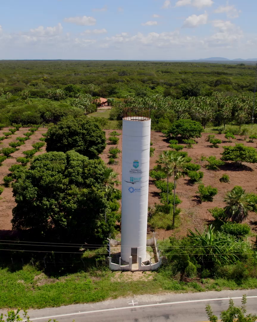Vista aérea de um tanque de água potável alto e cilíndrico, com o texto 'Sistema de água potável 24/7'. O tanque está rodeado por vegetação e terra, com detalhes de logotipos e texto adicional em sua estrutura.